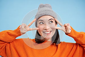 Close up portrait of cute korean girl in beanie, shows peace, vsign gesture, positive vibe, smiling and laughing, posing