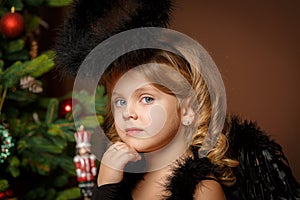 Close-up portrait of a cute little blonde girl with blue eyes in a black demon-demon costume against the background of a Christmas