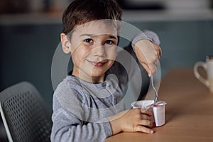 Close up of portrait of cute little boy eats delicious yogurt or cottage cheese from white container with spoon at table in dining