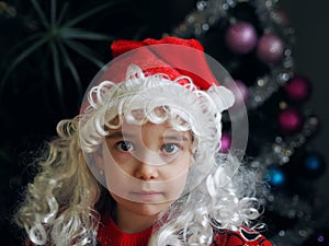 Close up portrait of cute little Christmas girl wearing red Santa hat and red sweater. Christmas tree background and defocused