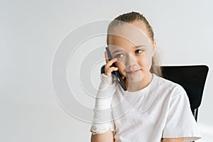 Close-up portrait of cute little girl with broken arm wrapped in plaster bandage talking smartphone, smiling looking