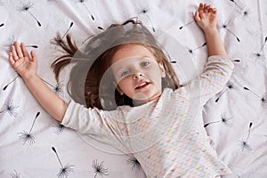 Close up portrait of cute little girl dreaming in bed, lying on white linen with dandelion and spreading arms, looking at camera,