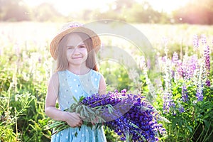 Close-up portrait of cute little girl in a hat with a bouquet of lupins in sunlight. Girl holding a bouquet of purple flowers in a
