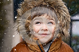 Close up portrait of cute little toddler boy in furry hood looking at snowfall