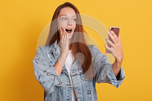 Close up portrait of cute lovely woman taking selfie, posing over yellow background, lady with open mouth holding her mobile phone