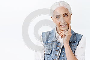 Close-up portrait cute, modern charming senior lady with combed grey hair in denim vest, slightly touching sking and
