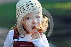 Close up portrait of cute one year old girl in a white knitted hat eating cookie on a stroll in the park