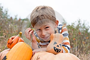 Close-up portrait of cute preschooler boy sitting in field near big pumpkins decorated for Halloween. October, outdoor. Harvesting