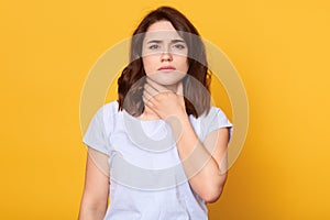 Close up portrait of cute sick young brunette woman wearing white casual t shirt, having sore throat, holding hand on her neck, photo