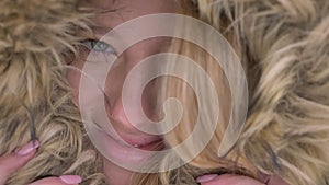 Close-up portrait of cute young caucasian girl hiding in fur hood prettily smiling into camera on winter street