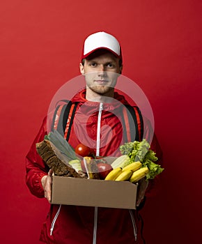 Close up portrait of delivery man in red uniform delivering food, groceries, vegetables, drinks in a paper box to a