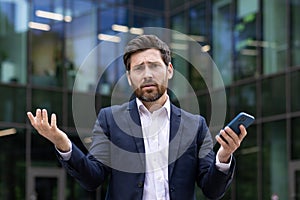 Close-up portrait of a disappointed young businessman standing in a suit near an office building, holding a phone and