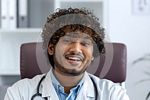 Close-up portrait of doctor inside clinic at workplace, man in white medical coat smiling looking at camera, satisfied