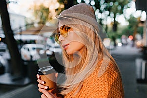 Close-up portrait of dreamy fair-haired girl waiting friend outdoor and drinking latte. Fascinating female model in