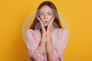 Close up portrait of emotional female being shocked to see something, opens mouth with surprisment and keepspalms near cheek,