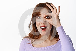 Close up portrait of excited stylish girl, looking through OK okay sign against eye, watching something awesome