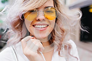 Close-up portrait of fair-haired laughing girl wearing trendy yellow sunglasses. Outdoor photo of happy blonde young