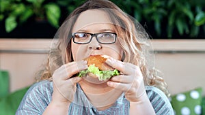 Close-up portrait of fat smiling woman enjoying biting fresh appetizing burger looking at camera