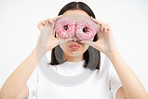 Close up portrait of funny asian girl looks through donnut holes, holding two glazed doughnuts and making cute faces photo