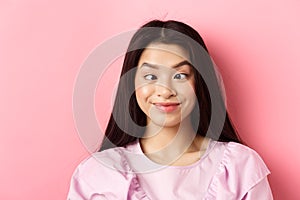 Close-up portrait of funny asian woman squinting eyes and making silly faces, standing against pink background