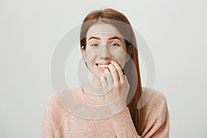 Close-up portrait of funny emotive caucasian ginger girl with bad habit, biting nails on fingers while looking at camera