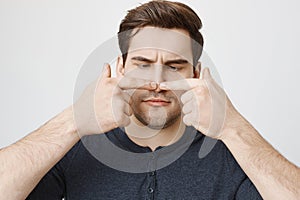 Close-up portrait of funny guy with cute haircut touching his nose with both index fingers and looking at it, standing