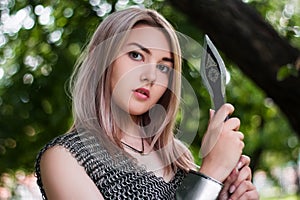 Close-up portrait of a girl in chain mail armor with a spear in her hands on the background of the forest