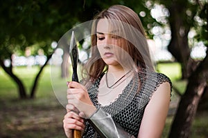 Close-up portrait of a girl in chain mail armor with a spear in her hands on the background of the forest