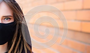 Close-up portrait of girl with dreadlocks and green-blue eyes, wearing medical flu mask and hooded sweater, looking straight, on