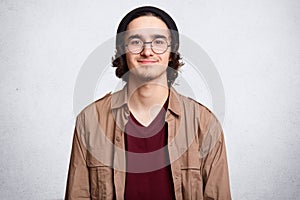 Close up portrait of good looking boy with curly hair, beard, wears round transparent glasses, models against white background.