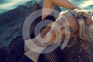 Close up portrait of gorgeous glam tanned blond woman wearing black swimsuit and summer tunic relaxing on the stones at the sea