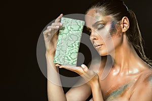 Close up portrait of gorgeous woman with closed eyes and artistic snakeskin make up holding green leather purse at her face
