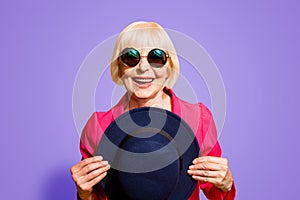 Close up portrait of granny in summer glasses holds a black hat in front of her isolated on vivid yellow background