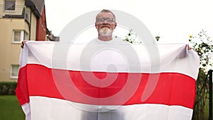 Close-up portrait of a gray-haired pensioner with glasses and a beard holding the red and white flag of Belarus. Protest