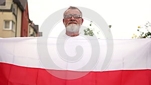 Close-up portrait of a gray-haired pensioner with glasses and a beard holding the red and white flag of Belarus. Protest