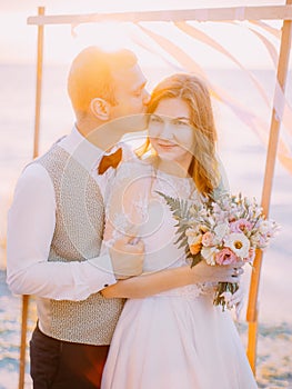 The close-up portrait of the groom kissing the bride on th head at the background of the sunset.