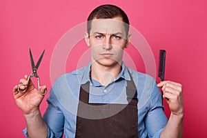 Close up portrait of handsome barber with serious facial expression, holds scissors, hair comb, ready for making hairstyle, looks