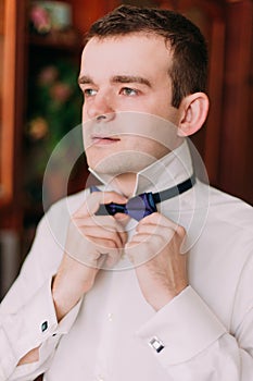 Close-up portrait of handsome businessman in blue suit putting on bow-tie indoors