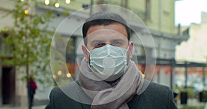 Close up portrait of handsome male in medical mask standing outdoors on street and looking at camera. Caucasian serious