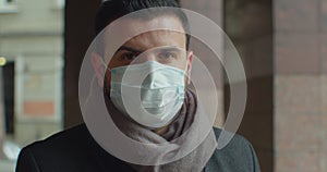 Close up portrait of handsome male in medical mask standing outdoors on street and looking at camera. Caucasian serious