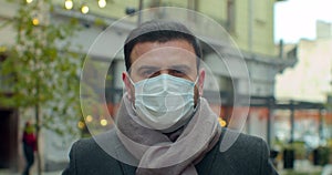 Close up portrait of handsome male in medical mask standing outdoors on street and looking at camera. Caucasian serious