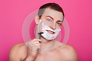 Close up portrait of handsome man with foam on face, shaving with razor in bathroom, looking smiling at camera, posing with bared