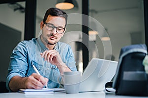 Close up portrait of handsome man working from home office taking reading and writing notes in note pad while working on laptop