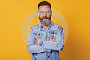 Close up portrait of handsome middle aged Caucasian man with beard, posing isolated on yellow background, wears denim jacket and