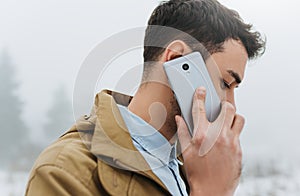 Close up portrait of handsome stylish young Caucasian man wearing trendy coat, looking down and talking on his mobile phone during