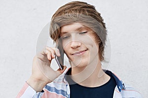 Close up portrait of handsome young boy with narrow dark eyes, stylish hairdo, dimple on cheek holding his mobile phone on ear cal