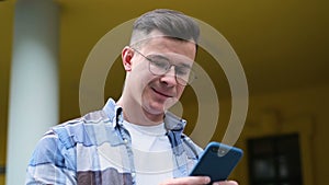 Close-up portrait of handsome young man using his smartphone scrolls through social media feed on device, giggles and