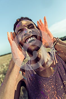 Happy african american man with colorful paint of face laughing with closed eyes at holi festival