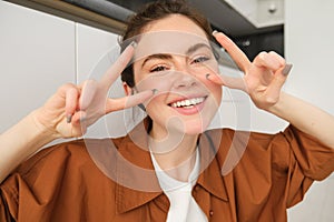 Close up portrait of happy, beautiful girl listens to music in wireless headphones, shows peace, v-sign gestures and