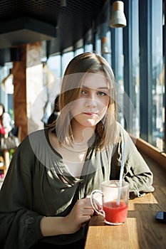 Close up portrait of happy beautiful teen student girl with a glass mug straw fruit tea at street cafe sitting near big window bit